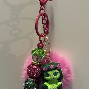 May include: A pink keychain with a cartoon character, a pink fluffy pom-pom, and decorative beads. The keychain has a lobster clasp and a key ring. The beads are green, pink, and dark blue with gold accents.