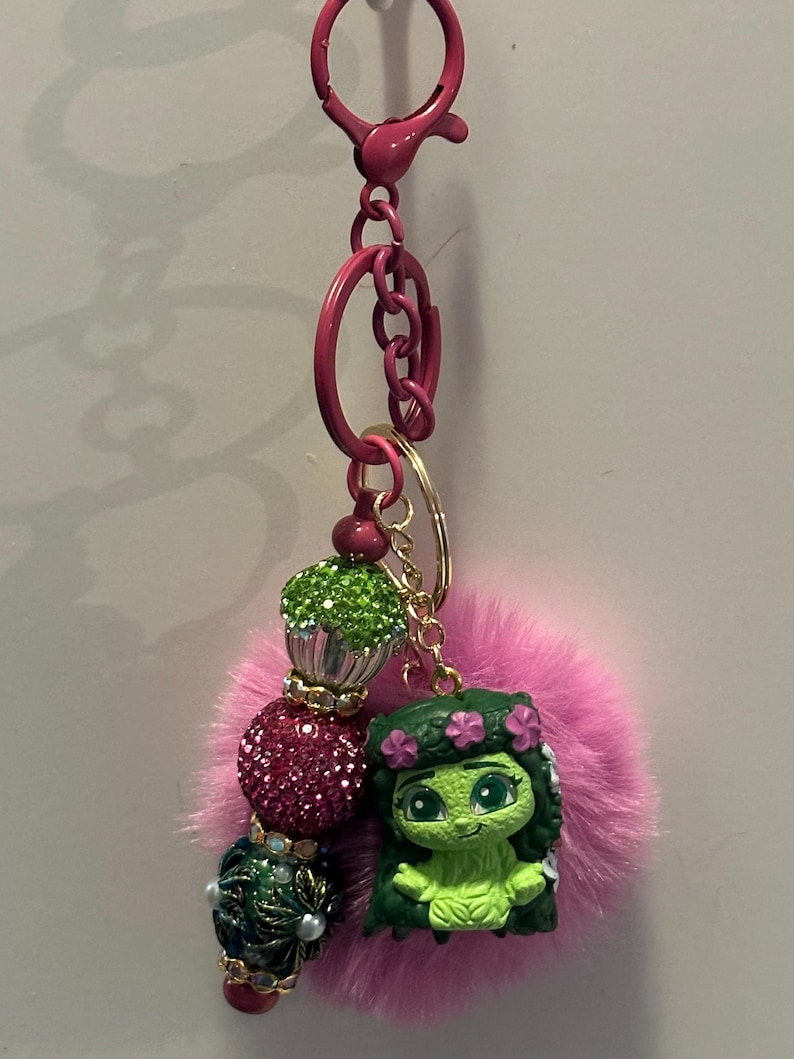 May include: A pink keychain with a cartoon character, a pink fluffy pom-pom, and decorative beads. The keychain has a lobster clasp and a key ring. The beads are green, pink, and dark blue with gold accents.