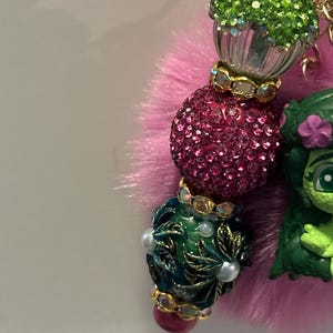 May include: A close-up of a beaded charm with a pink fluffy base. The charm features a green cupcake bead, a sparkly pink bead, and a dark green bead with white pearls. A green cartoon character is also attached.