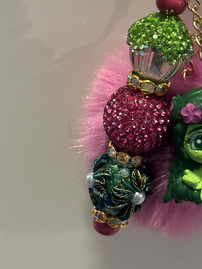 May include: A close-up of a beaded charm with a pink fluffy base. The charm features a green cupcake bead, a sparkly pink bead, and a dark green bead with white pearls. A green cartoon character is also attached.