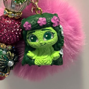 May include: A green figurine of a cartoon character with a flower crown, attached to a pink fluffy pom-pom keychain. The keychain also features decorative beads in shades of green, pink, and gold.