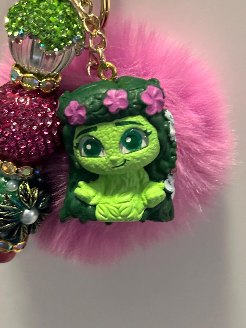 May include: A green figurine of a cartoon character with a flower crown, attached to a pink fluffy pom-pom keychain. The keychain also features decorative beads in shades of green, pink, and gold.