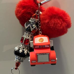 Puede incluir: Un llavero rojo con un dije de cami&oacute;n Mack de dibujos animados, un dije de cuentas con pedrer&iacute;a roja y negra, y dos pompones rojos y esponjosos. El llavero est&aacute; sujeto a un mosquet&oacute;n rojo.