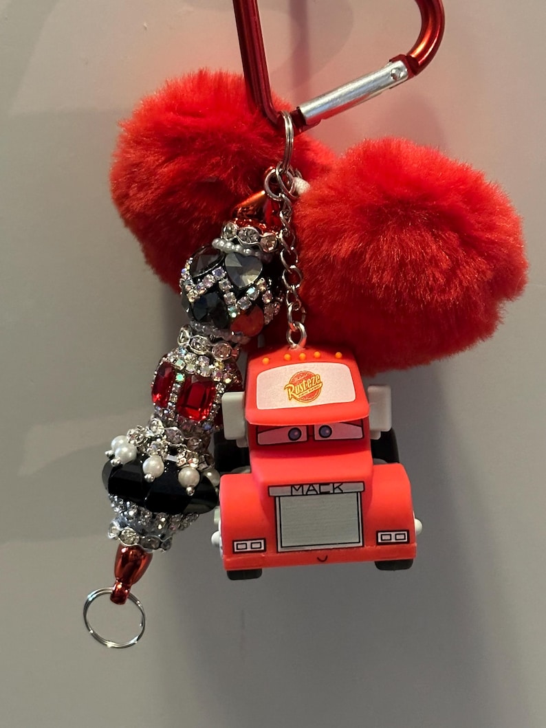 Puede incluir: Un llavero rojo con un dije de cami&oacute;n Mack de dibujos animados, un dije de cuentas con pedrer&iacute;a roja y negra, y dos pompones rojos y esponjosos. El llavero est&aacute; sujeto a un mosquet&oacute;n rojo.