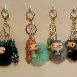 May include: Four keychains with gold-colored hardware, each featuring a small character figure and a fluffy pom-pom. The figures are from a popular fantasy series. The keychains are hanging against a white background.