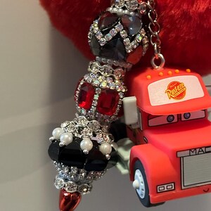 Puede incluir: Un llavero con cuentas con un dije en forma de coraz&oacute;n rojo. El llavero presenta una serie de cuentas decorativas en negro, rojo y detalles de cristal transparente. Un peque&ntilde;o cami&oacute;n de juguete rojo con la palabra "MAC" est&aacute; adjunto al llavero.