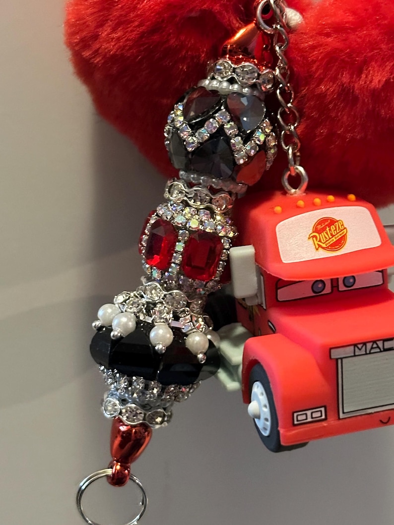 Puede incluir: Un llavero con cuentas con un dije en forma de coraz&oacute;n rojo. El llavero presenta una serie de cuentas decorativas en negro, rojo y detalles de cristal transparente. Un peque&ntilde;o cami&oacute;n de juguete rojo con la palabra "MAC" est&aacute; adjunto al llavero.