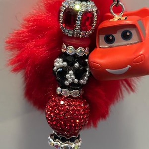 K&ouml;nnte beinhalten: Ein rot-schwarzer Perlen-Schl&uuml;sselanh&auml;nger mit einem roten, flauschigen Akzent und einem kleinen orangefarbenen Spielzeugauto. Die Perlen sind mit Strasssteinen und Kristallen verziert. Der Schl&uuml;sselanh&auml;nger ist an einer silbernen Kette befestigt.