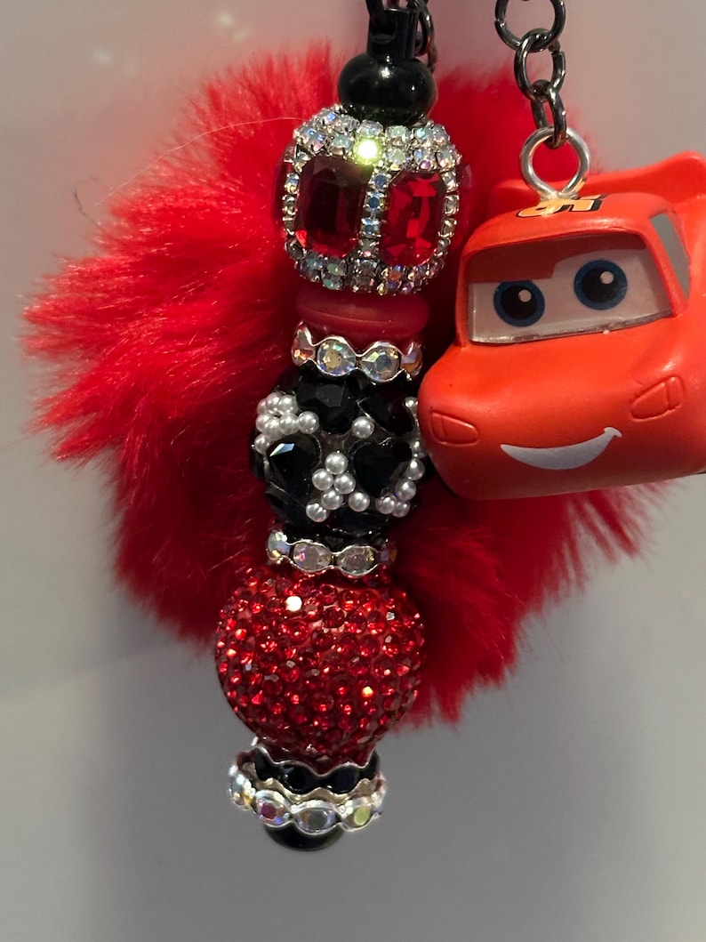 K&ouml;nnte beinhalten: Ein rot-schwarzer Perlen-Schl&uuml;sselanh&auml;nger mit einem roten, flauschigen Akzent und einem kleinen orangefarbenen Spielzeugauto. Die Perlen sind mit Strasssteinen und Kristallen verziert. Der Schl&uuml;sselanh&auml;nger ist an einer silbernen Kette befestigt.