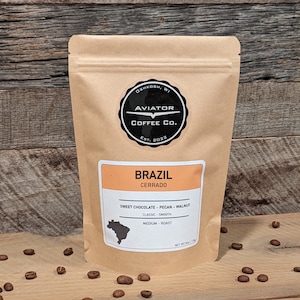 May include: A brown paper bag of Brazil Cerrado coffee with the Aviator Coffee Co. logo. The label indicates sweet chocolate, pecan, and walnut flavors. The bag is on a wooden surface with scattered coffee beans.
