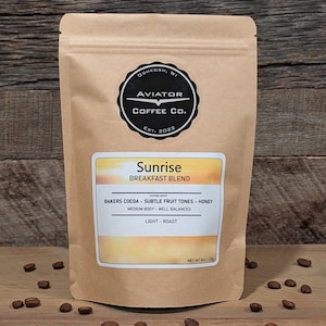May include: A brown paper bag with a white label that says "Sunrise Breakfast Blend" and lists the coffee's tasting notes: "Bakers Cocoa - Subtle Fruit Tones - Honey", "Medium Body - Well Balanced", and "Light Roast". The bag is labeled "Aviator Coffee Co. Est. 2022" and is sitting on a wooden surface with coffee beans scattered around it.