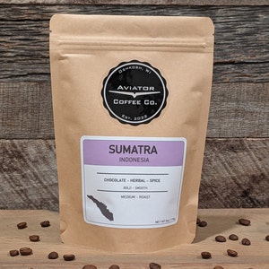 May include: A brown paper bag of Sumatra Indonesia coffee beans from Aviator Coffee Co. The bag has a black and white circular logo and a purple label with flavor notes. The coffee is described as chocolate, herbal, and spice, with a medium roast.