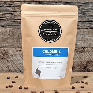 May include: A brown paper bag with a label that says "Colombia Bucaramanga" and has a map of Colombia on it. The label also says "Almond - Cocoa - Hint of Vanilla", "Medium Body - Silky", and "Medium Roast". The bag is from Aviator Coffee Co. and is on a wooden surface with coffee beans scattered around it.