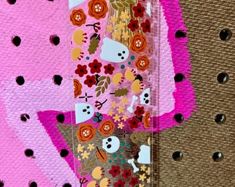 May include: A clear rectangular bookmark featuring a pattern of white ghosts, white skulls, white bones, red flowers, orange flowers, and green leaves. The bookmark is topped with a metallic gold tassel made of thin, shimmering corded strands.