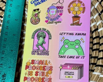 May include: A sheet of colourful stickers with various designs and positive messages. The stickers feature frogs, flowers, and a retro game console. The text on the stickers includes "You can always start again", "Nothing to prove, just want to groove", "Small progress is still progress", "Give yourself time to grow and heal", "Letting karma take care of it", and "Don't let it all build up".