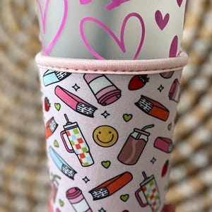 May include: A clear plastic cold cup with a lid, decorated with pink hearts. The cup is inside a pink sleeve with a pattern of books, drinks, strawberries, and smiley faces. The sleeve has a pink trim.