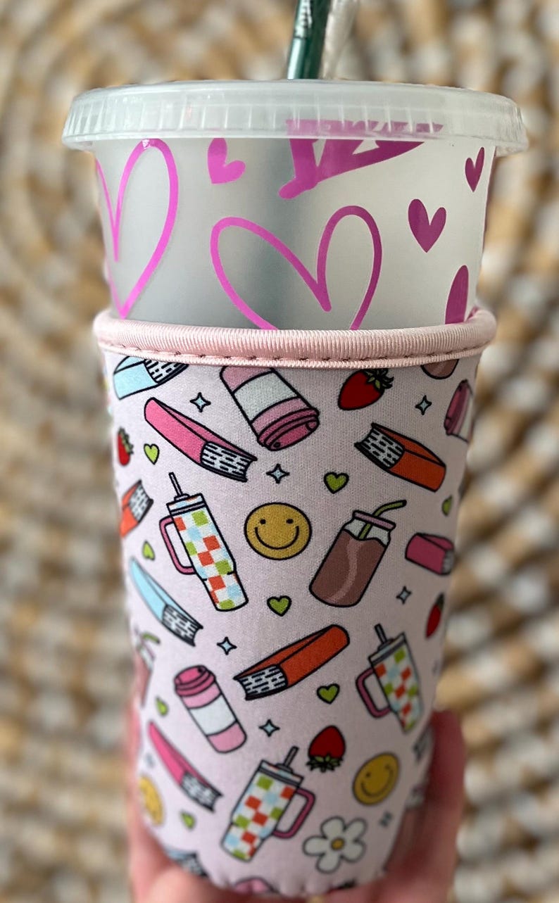 May include: A clear plastic cold cup with a lid, decorated with pink hearts. The cup is inside a pink sleeve with a pattern of books, drinks, strawberries, and smiley faces. The sleeve has a pink trim.