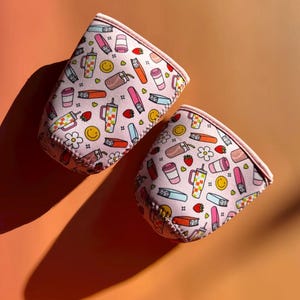 May include: Two pink drink sleeves with a repeating pattern of coffee cups, lipsticks, strawberries, and smiley faces. The sleeves are made of a soft material and have a pink trim. The background is a solid orange colour.