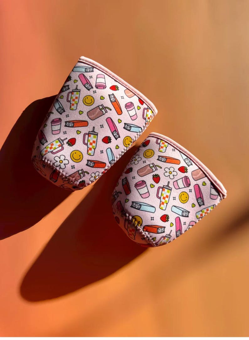 May include: Two pink drink sleeves with a repeating pattern of coffee cups, lipsticks, strawberries, and smiley faces. The sleeves are made of a soft material and have a pink trim. The background is a solid orange colour.