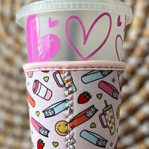 May include: A clear plastic tumbler with a pink heart design and a green straw. The tumbler is in a pink patterned sleeve with books, strawberries, and smiley faces. The sleeve is held by a hand.