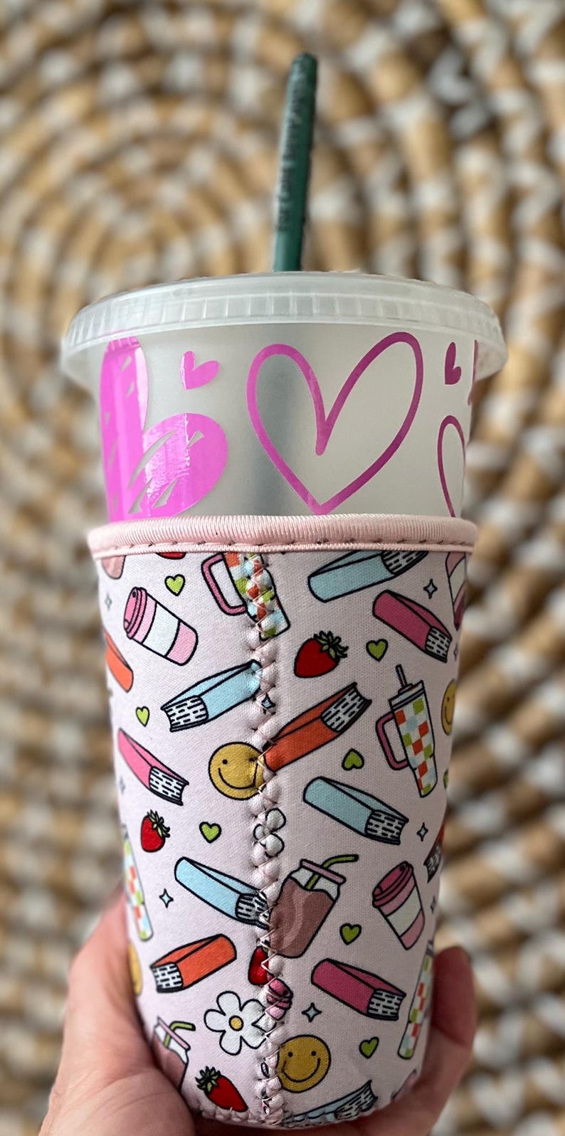 May include: A clear plastic tumbler with a pink heart design and a green straw. The tumbler is in a pink patterned sleeve with books, strawberries, and smiley faces. The sleeve is held by a hand.