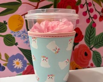 May include: A light blue neoprene cup sleeve featuring a repeating pattern of small white ghosts wearing colourful flower crowns. The sleeve is fitted around a clear plastic cup with a clear lid, containing pink tissue paper inside the cup.