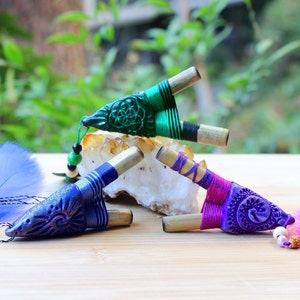 May include: Three handmade wooden pipes with colorful designs and feathers. The pipes are decorated with green, blue, and purple string and have a geometric design.