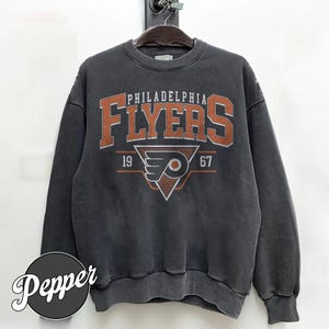 Könnte beinhalten: Ein anthrazitfarbenes Sweatshirt mit Rundhalsausschnitt und dem Philadelphia Flyers-Logo in Orange, Weiß und Schwarz. Der Aufdruck zeigt den Schriftzug Philadelphia Flyers, das Jahr 1967 und ein geflügeltes P-Emblem in einem Dreieck.