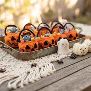 May include: A collection of small orange crocheted pumpkin baskets with black yarn handles and stitched jack-o'-lantern faces, each filled with assorted candies. Beside the metal tray sits a white ceramic ghost and two small white pumpkins.