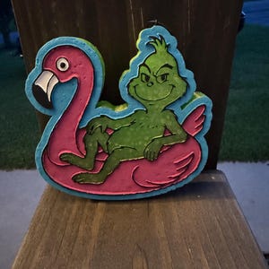 May include: A decorative wall hanging featuring a green, mischievous character lounging inside a bright pink flamingo pool float. The piece has a light blue border, a black outline, and a metal loop at the top for hanging. The design is colorful and flat.
