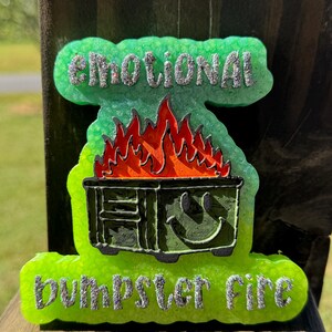 May include: A bright neon green resin plaque with a textured surface. The text EMOTIONAL is at the top and dumpster fire is at the bottom in silver glitter. A dark green dumpster with a smiley face and orange flames is in the center.