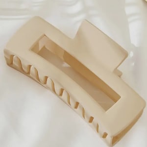 May include: A beige rectangular hair claw clip. The clip has a matte finish and a simple, modern design. The clip has a rectangular opening in the center and wavy teeth on one side for grip. A stylish hair accessory.