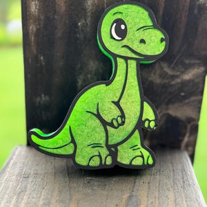 May include: A bright green, flat, cartoon-style dinosaur figure with a long neck and a rounded head. The dinosaur has a black outline, large white eyes with black pupils, and a textured surface finish, standing in a side-facing profile position.