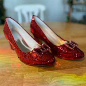 May include: A pair of red sequined low-heeled shoes with a bow detail. The shoes have a pointed toe and a small heel. The sequins give the shoes a shiny appearance. The shoes are displayed on a wooden surface.