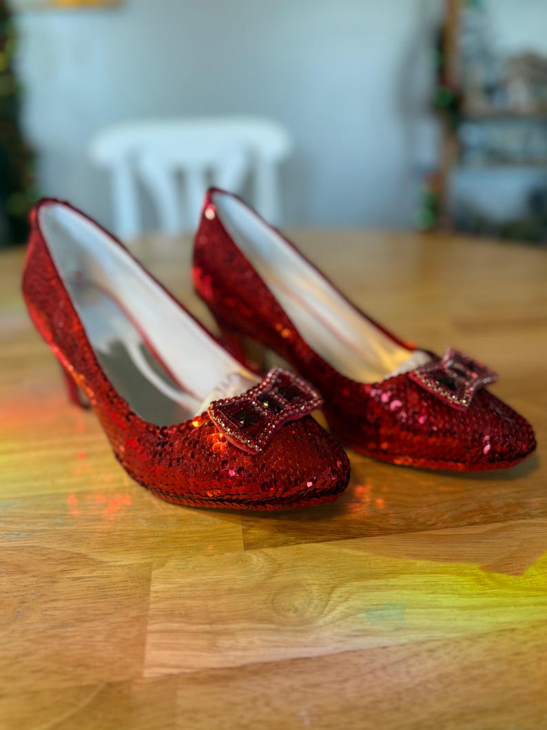 Pu&ograve; includere: Un paio di scarpe rosse con paillettes e tacco basso con dettaglio a fiocco. Le scarpe hanno la punta e un tacco piccolo. Le paillettes conferiscono alle scarpe un aspetto lucido. Le scarpe sono esposte su una superficie di legno.