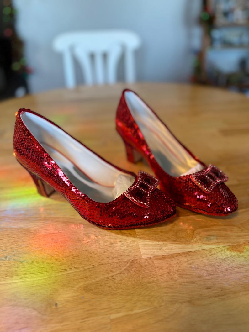 Pu&ograve; includere: Un paio di d&eacute;collet&eacute; rosse con paillettes e un fiocco sulla punta. Le scarpe hanno un tacco basso e un interno bianco. Le scarpe sono appoggiate su una superficie di legno.