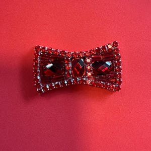 May include: A red bow-shaped brooch with a sparkling design. The brooch features a central rectangular gem and is surrounded by smaller red and clear crystals. The background is a solid red color.