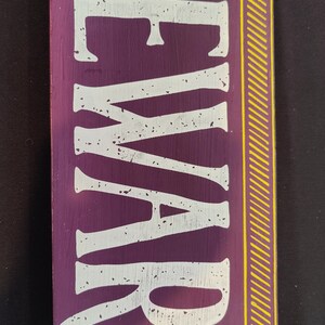 May include: Purple wooden sign with yellow stripes on the right side. The sign says "BEWARE" in white distressed lettering.