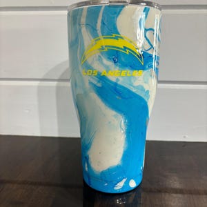 May include: A stainless steel tumbler with a blue and white marbled design. The tumbler features a yellow and blue Los Angeles Chargers logo and the words "LOS ANGELES" in yellow.