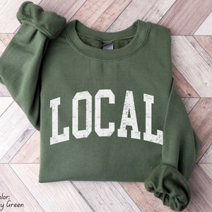 May include: A green sweatshirt with the word "LOCAL" printed in white on the front. The sweatshirt has a distressed, vintage look.