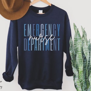 May include: Navy blue sweatshirt with the words "EMERGENCY nurse DEPARTMENT" in blue and white lettering. The sweatshirt is hanging on a wooden hanger.