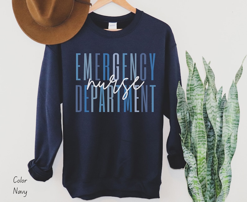 May include: Navy blue sweatshirt with the words "EMERGENCY nurse DEPARTMENT" in blue and white lettering. The sweatshirt is hanging on a wooden hanger.