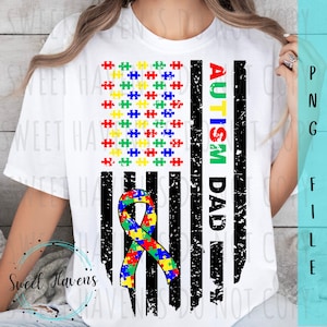 Op de afbeelding: Wit T-shirt met een verweerd Amerikaans vlagontwerp. De vlag is zwart-wit met een blauw, groen, geel en rood puzzelstukje lint in de linkerbenedenhoek. De woorden "Autism Dad" staan verticaal aan de rechterkant van de vlag.