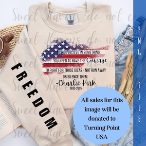 Op de afbeelding: Een beige sweatshirt met het woord "FREEDOM" in het zwart gedrukt op de linkermouw. De voorkant heeft een Amerikaans vlagontwerp en een citaat van Charlie Kirk. Een blauwe cirkel geeft aan dat de verkoop wordt gedoneerd aan Turning Point USA.