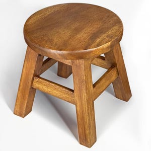 Könnte beinhalten: Ein kleiner, runder Holzschemel mit natürlicher Oberfläche. Der Hocker hat eine kreisförmige Sitzfläche und vier stabile, abgewinkelte Beine, die durch horizontale Streben verbunden sind. Die Holzmaserung ist sichtbar und verleiht ihm ein rustikales Aussehen.