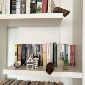 May include: A white bookshelf with two shelves, filled with books. The top shelf has a small potted plant and a wooden figurine of a cat. The bottom shelf has a small wooden house figurine, a clock, and a potted plant. The books on the shelves are mostly hardcover and have a variety of titles.