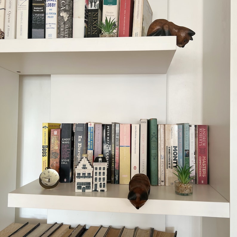 May include: A white bookshelf with two shelves, filled with books. The top shelf has a small potted plant and a wooden figurine of a cat. The bottom shelf has a small wooden house figurine, a clock, and a potted plant. The books on the shelves are mostly hardcover and have a variety of titles.