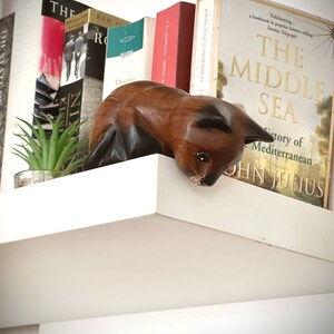 May include: A wooden cat figurine with brown and black fur, perched on a white shelf with a row of books behind it. The books have titles like "The Middle Sea" and "VS Naipaul India".
