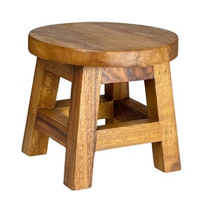 May include: A small, round wooden stool with a natural finish. The stool has a circular seat and four sturdy, square legs connected by a lower frame. The wood grain is visible, giving it a rustic appearance. The stool is approximately 30 cm in height.