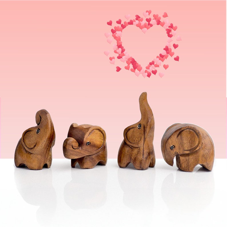 May include: Four wooden elephant figurines stand on a white surface, facing different directions. The elephants are carved in a simple style with brown wood. A pink background with a heart shape made of pink hearts is in the background.